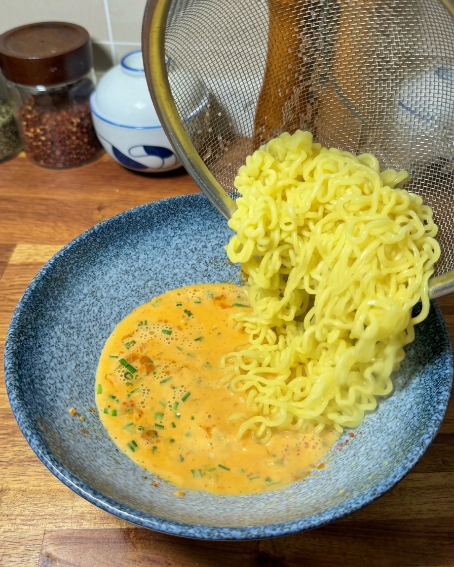 Fideos ramen amarillos y ondulados deslizándose desde un colador metálico hacia un bol con un rico caldo naranja.