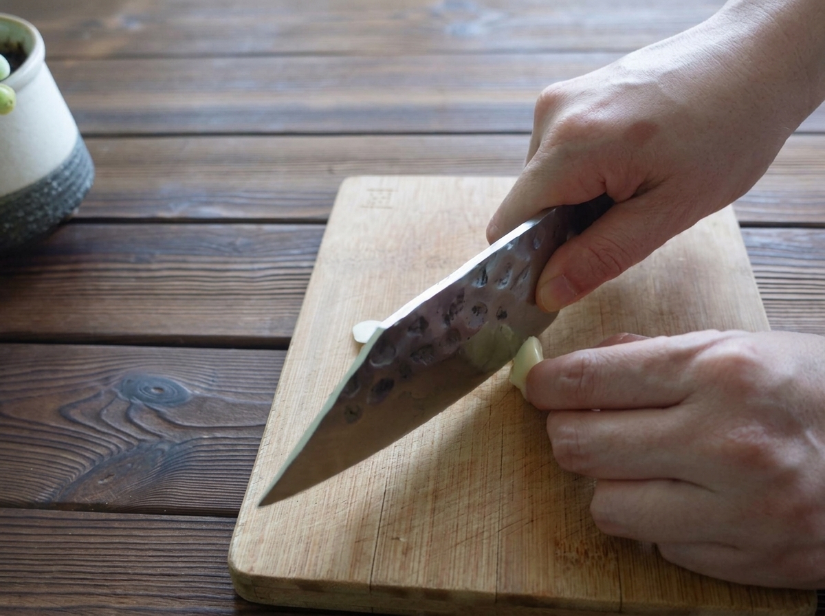 Manos cortando un diente de ajo en láminas finas sobre una tabla de madera.