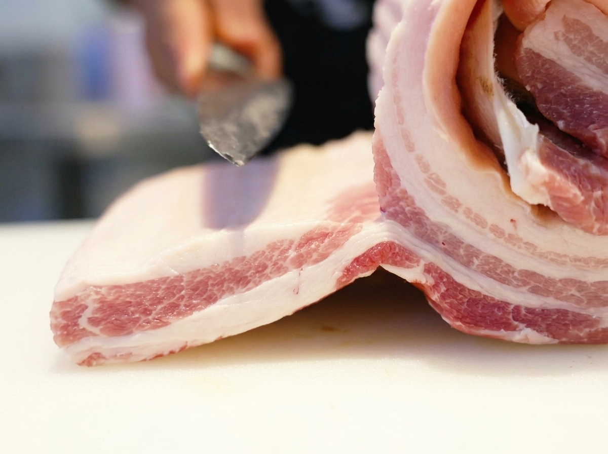 Cuchillo afilado cortando un bloque crudo de panceta de cerdo en una tabla blanca.