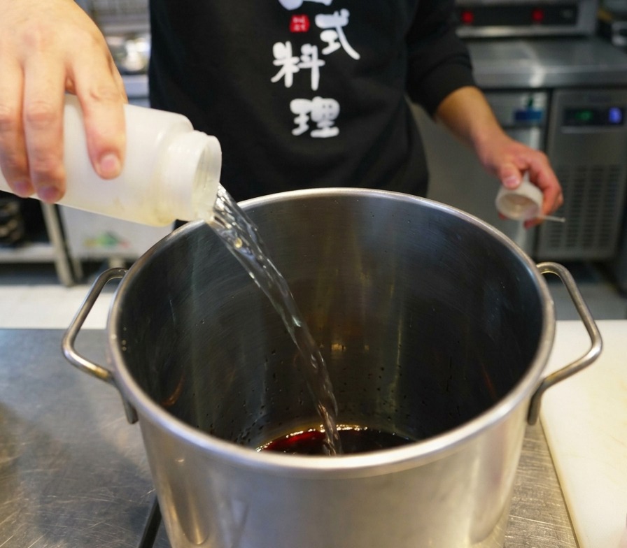 Persona vertiendo un líquido transparente desde un biberón de cocina a una olla de acero inoxidable.