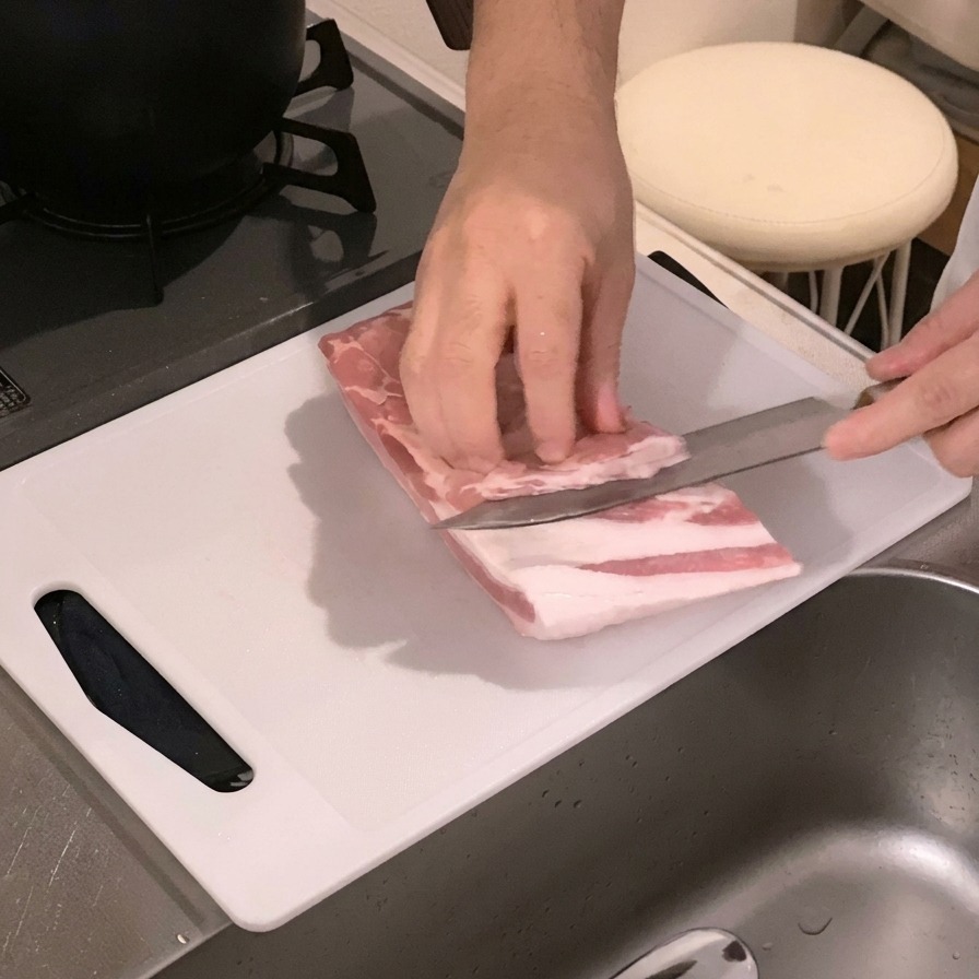 Manos marcando el interior de un bloque crudo de panceta de cerdo con un cuchillo sobre una tabla de cortar blanca.