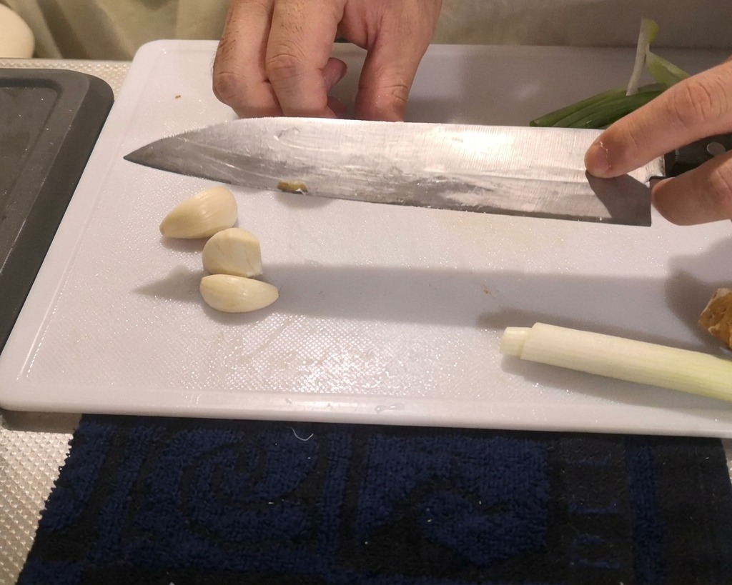 Una persona presionando la hoja plana de un cuchillo de cocina grande sobre dientes de ajo pelados para aplastarlos en una tabla de cortar.