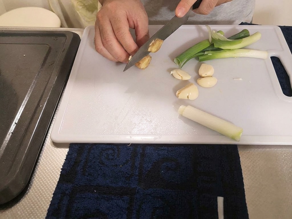 Un cocinero cortando un trozo de jengibre fresco en trozos gruesos sobre una tabla de cortar con cebolletas y ajo cerca.
