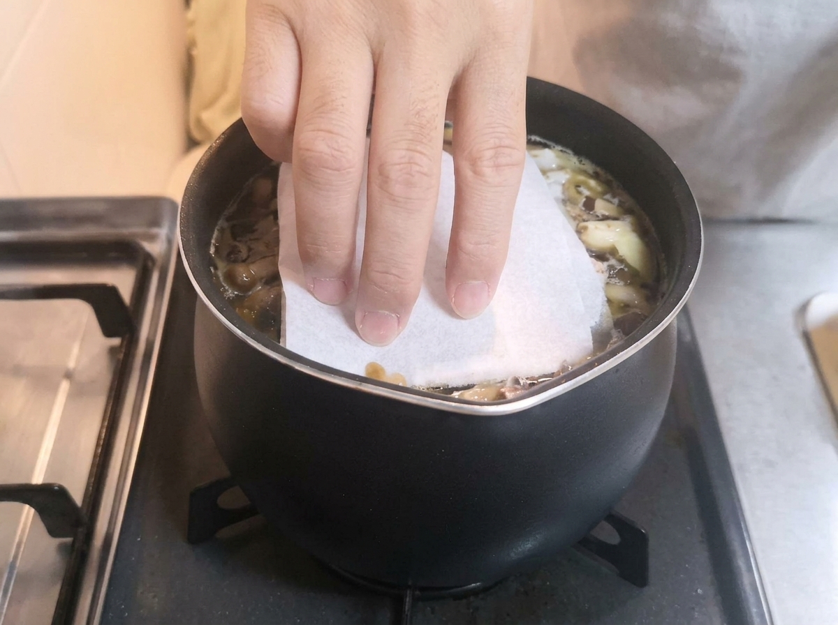 Una mano colocando una toalla de papel blanca sobre una olla a fuego lento con caldo oscuro y aromáticos.