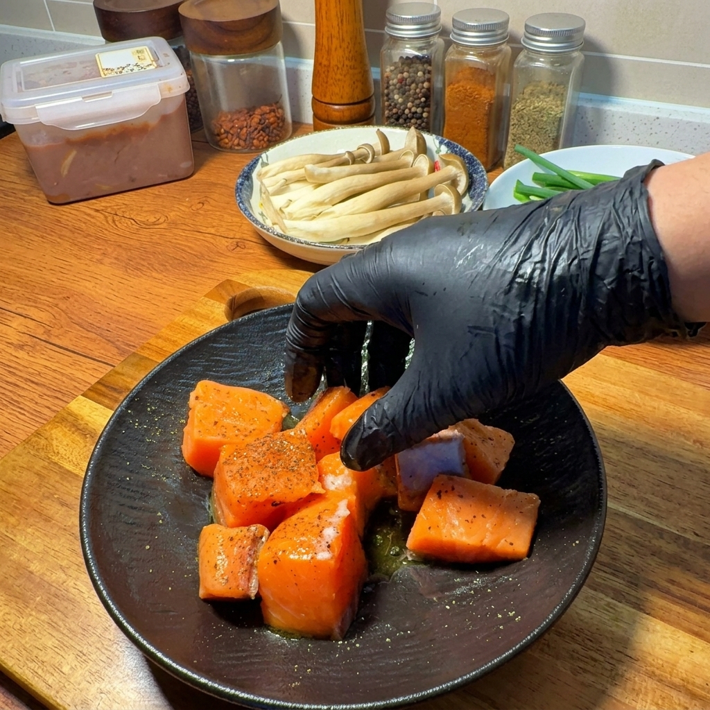 Una mano con guante mezclando trozos de salmón con especias y aceite en un plato oscuro.