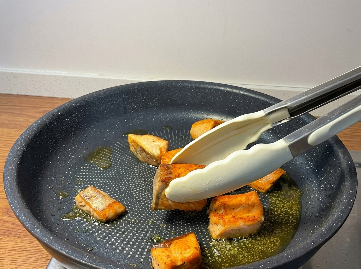 Pinzas dando la vuelta a trozos de salmón dorados en una sartén antiadherente.