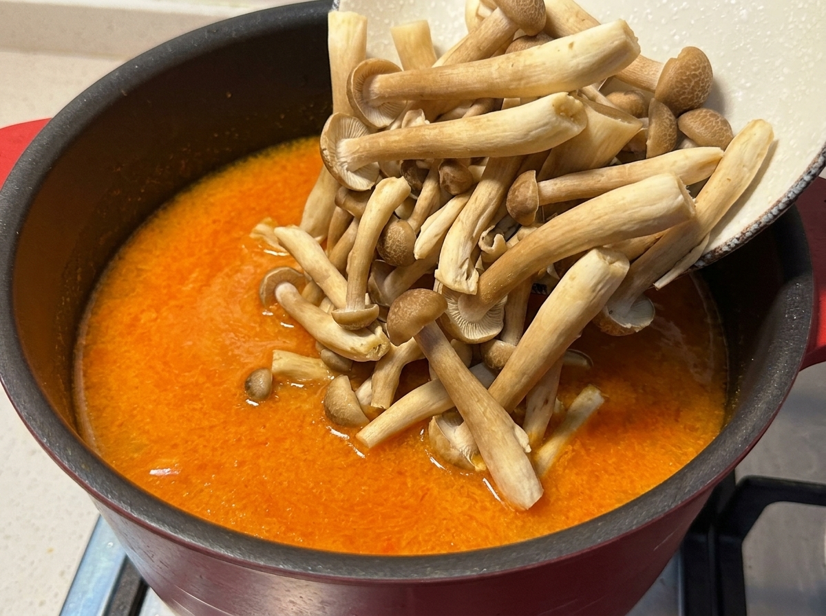 Echando setas shimeji frescas desde un bol a una olla con caldo de miso naranja hirviendo suavemente.