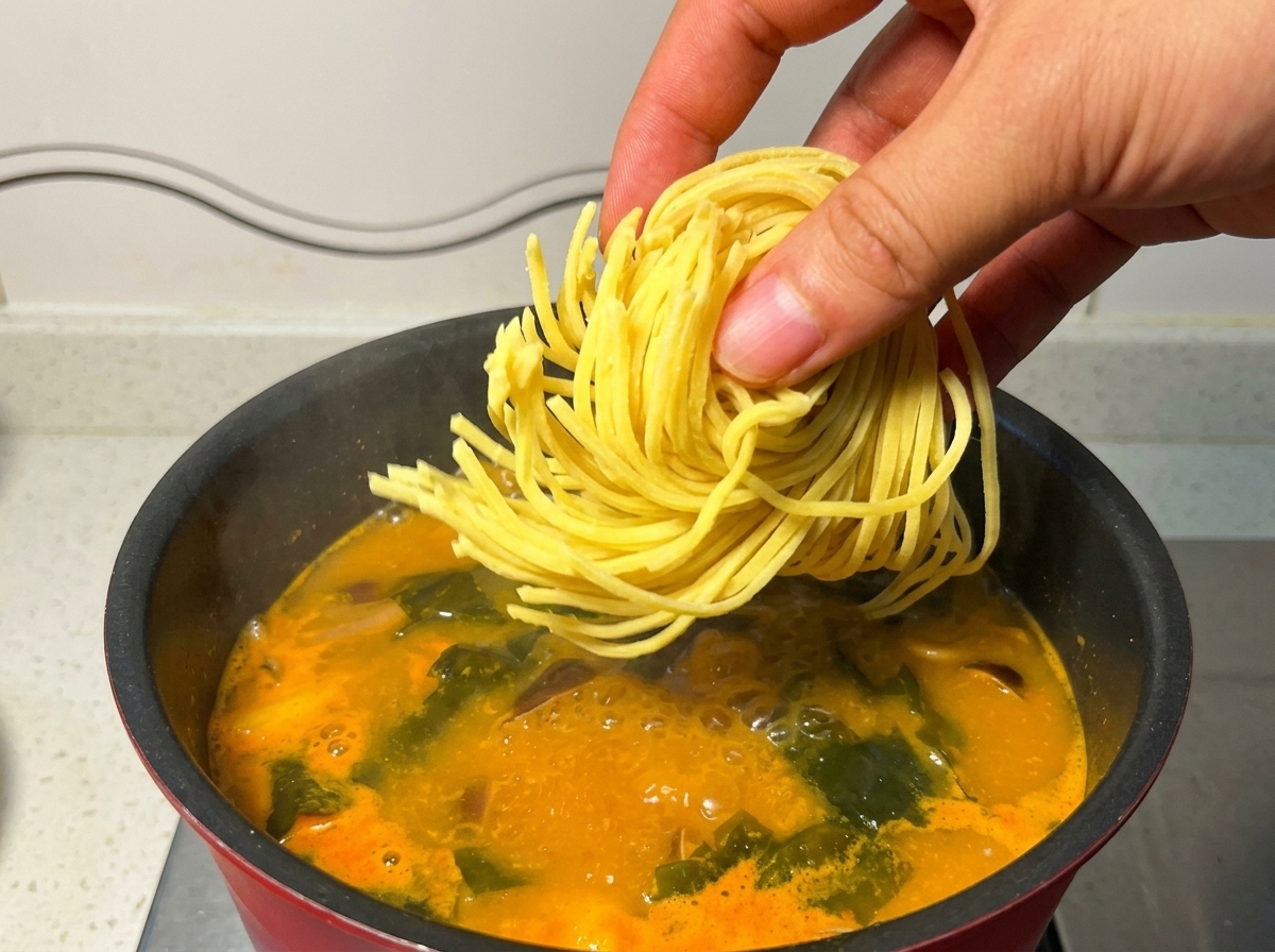 Una mano soltando un nido de fideos ramen amarillos frescos en una olla con sopa naranja hirviendo.