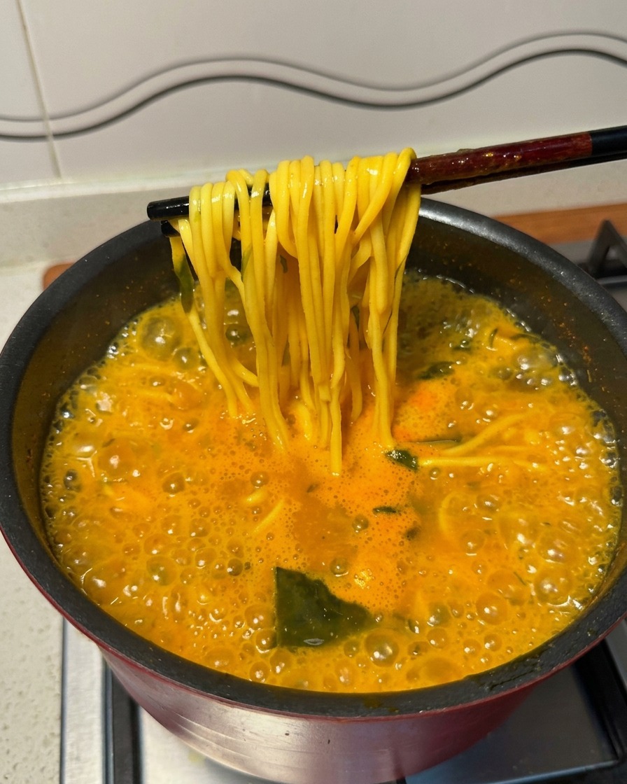 Palillos chinos negros levantando fideos ramen amarillos de una olla de sopa picante hirviendo con fuerza.