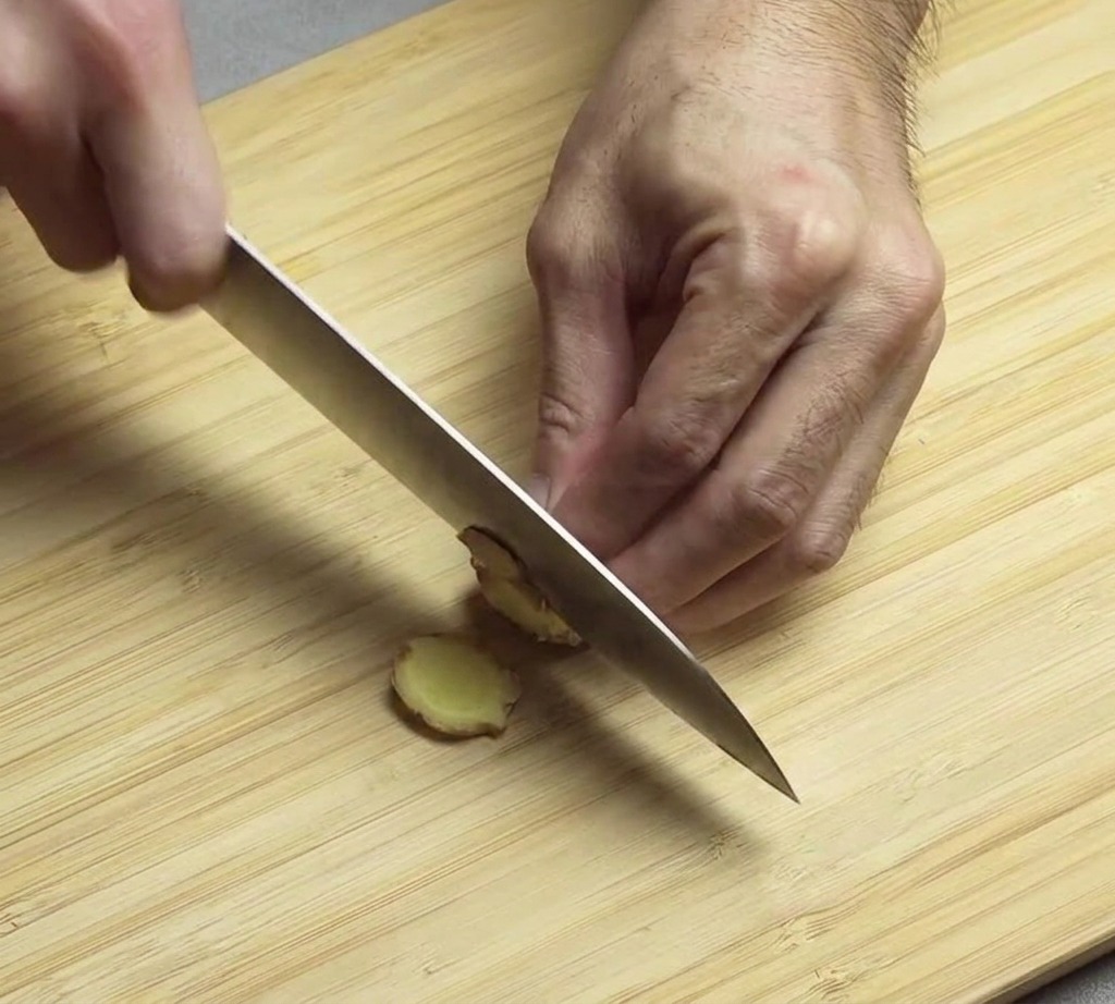 Manos usando un cuchillo de chef para picar finamente una pieza de jengibre fresco sobre una tabla de bambú.