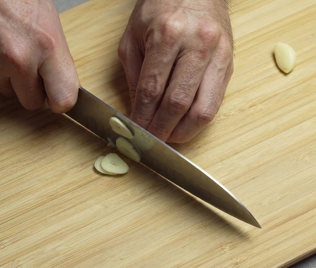 Manos picando finamente dientes de ajo pelados en una tabla de cortar de madera.