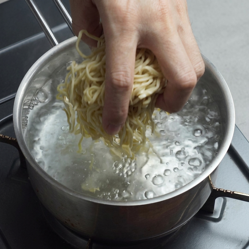 Una mano dejando caer fideos ramen frescos de color amarillo en una olla con agua hirviendo.