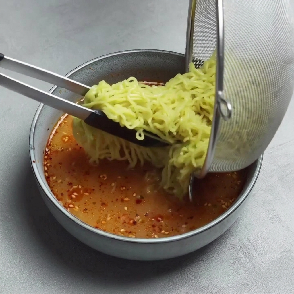 Pinzas transfiriendo fideos ramen cocidos desde un colador a un cuenco con caldo rojo picante.