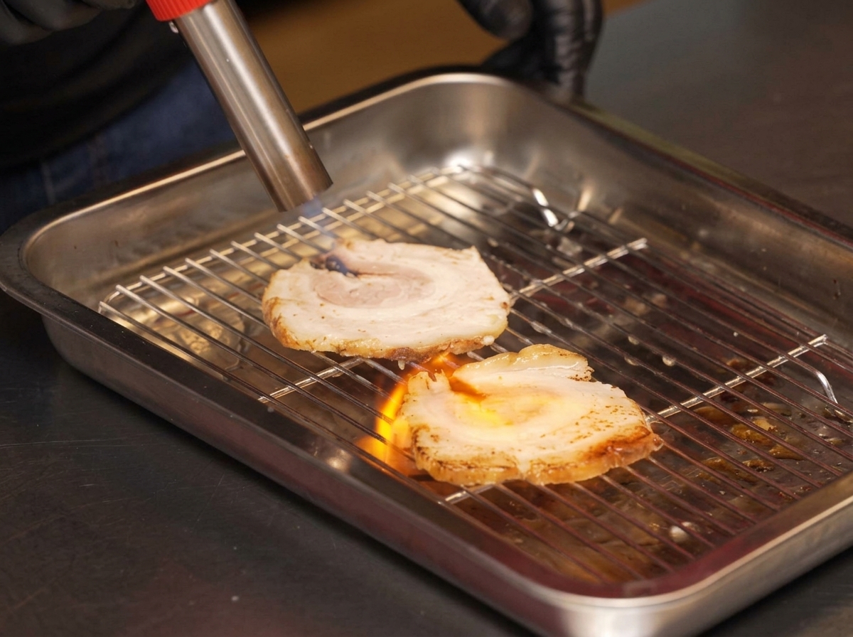 Un chef usando un soplete para brasear rodajas de chashu sobre una rejilla metálica.