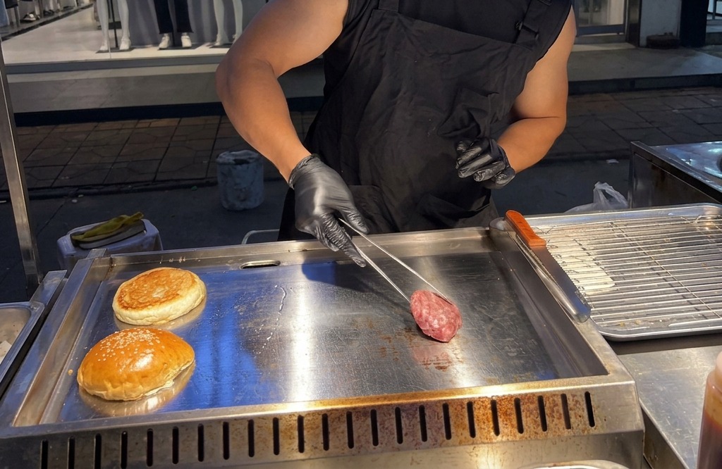 Una persona colocando una bola redonda de carne de res molida cruda sobre una plancha de metal humeante usando pinzas.