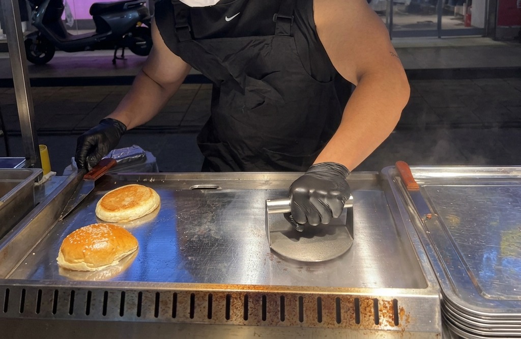 Una persona usando una prensa de metal pesada para aplastar firmemente una bola de carne de res sobre una plancha caliente.