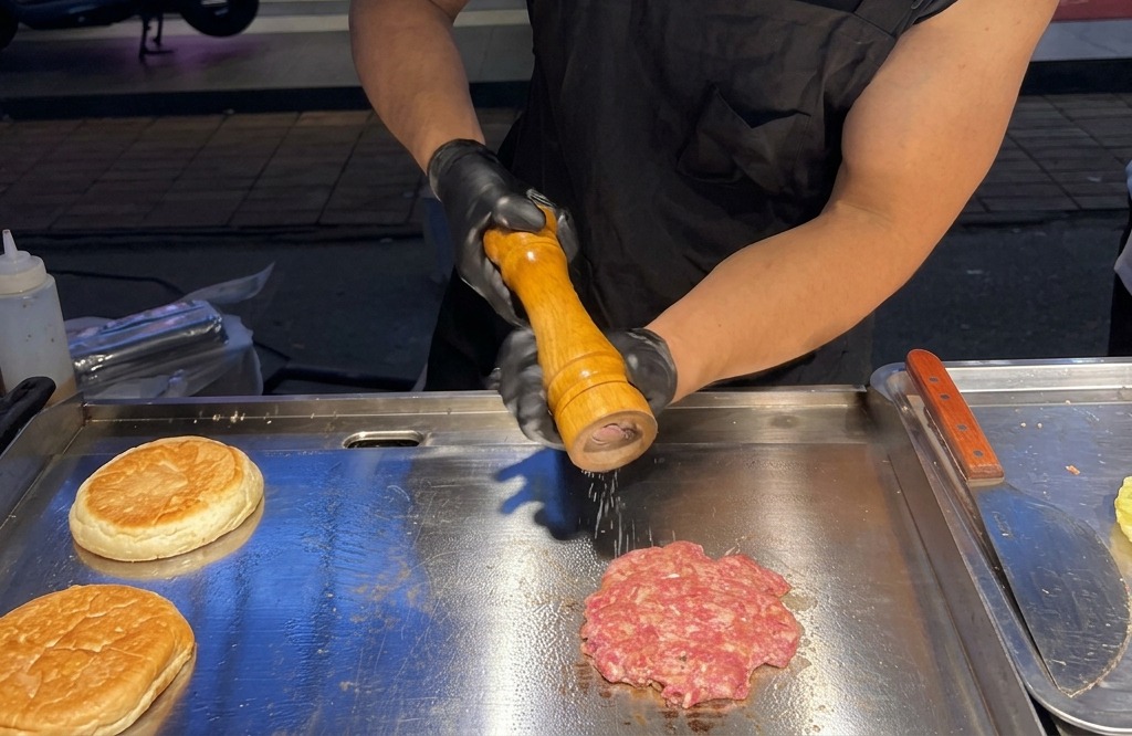 Moliendo sal marina y pimienta negra fresca sobre una hamburguesa de carne aplastada que chisporrotea en una plancha.