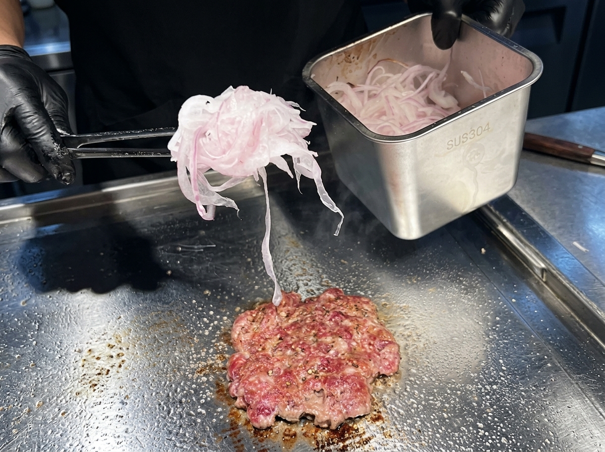 Un puñado de cebolla roja cortada muy fina siendo colocada sobre una hamburguesa de carne en una plancha caliente.