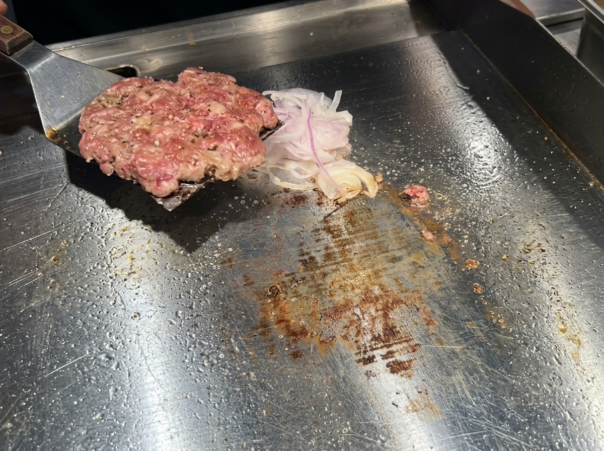 Una espátula de metal dando la vuelta a una hamburguesa de carne bien dorada sobre un montón de cebollas en una plancha.