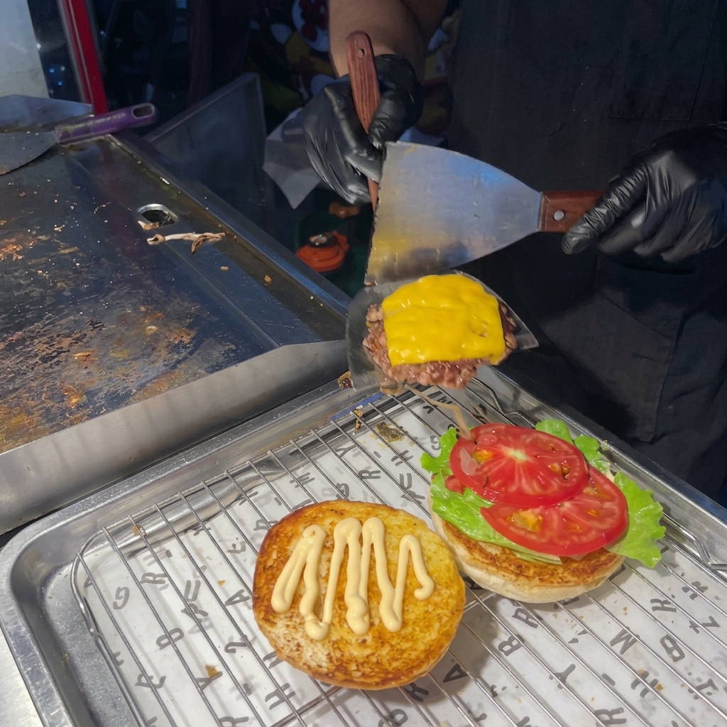 Una persona usando una espátula para colocar una hamburguesa cocinada con queso fundido sobre un pan con lechuga y tomate.