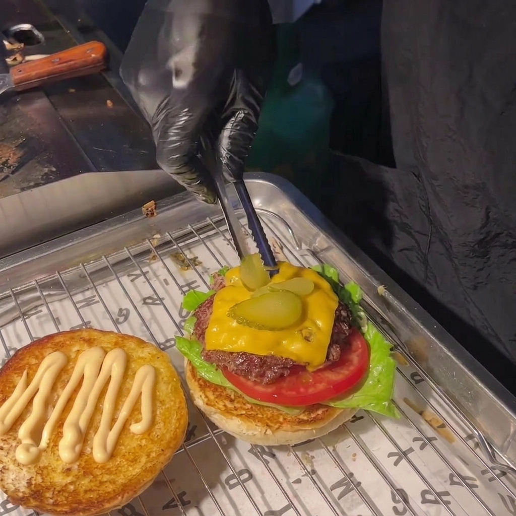 Una persona usando pinzas para colocar dos rodajas de pepinillo sobre el queso fundido de una hamburguesa.