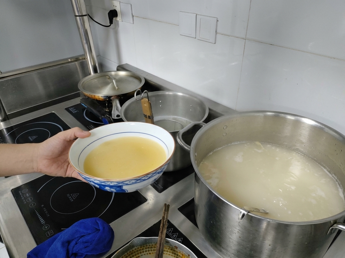 Una mano sosteniendo un bol con base de miso amarillo junto a una olla con caldo blanco hirviendo.