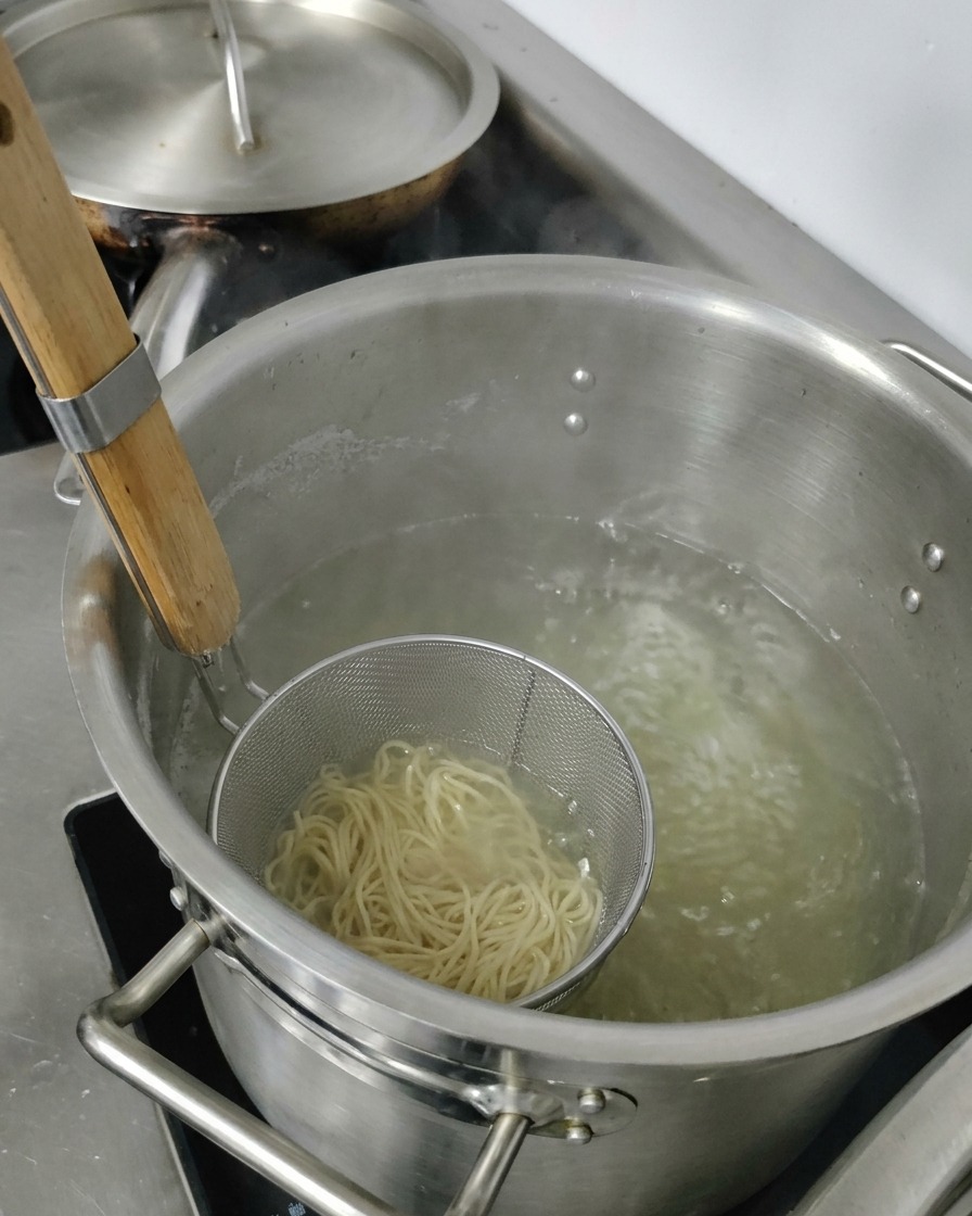 Levantando un colador metálico con fideos ramen cocidos de una olla con agua caliente.