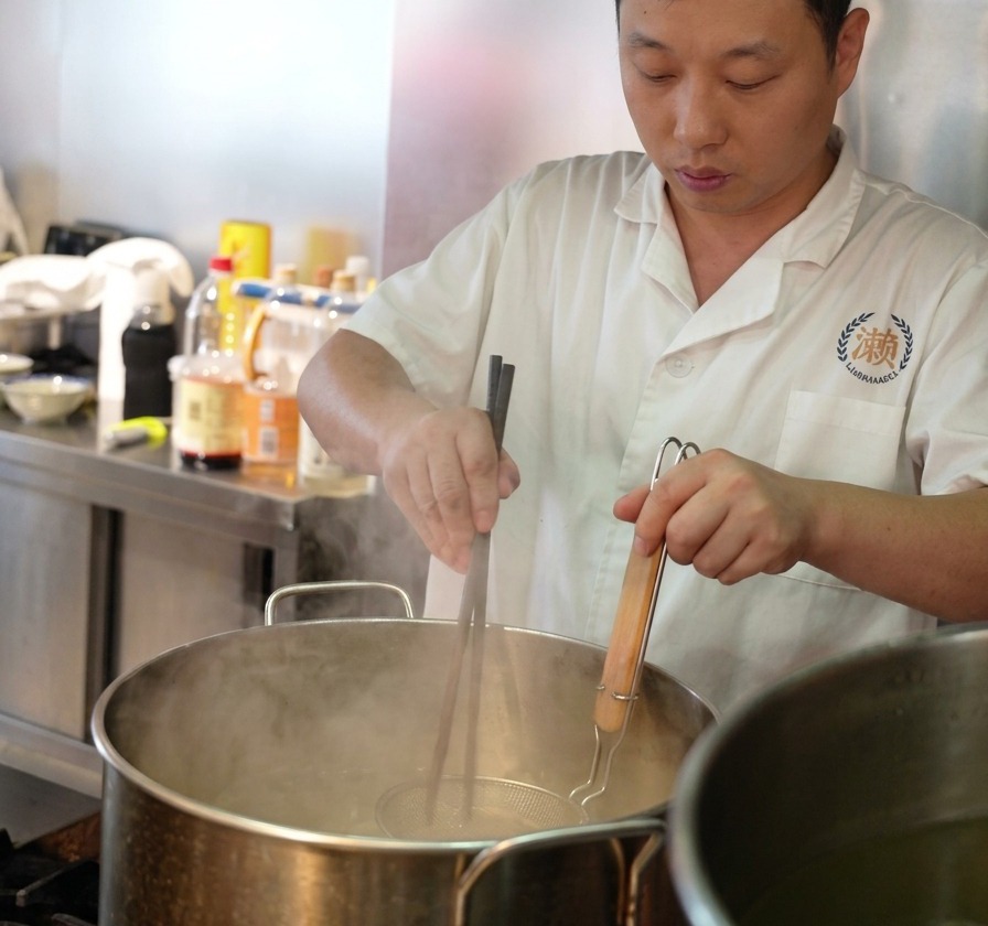 Un chef usa palillos largos para remover fideos frescos dentro de un colador metálico sumergido en agua hirviendo.