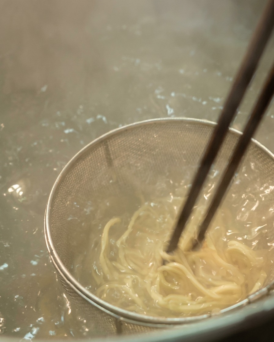 Primer plano de fideos ramen claros removiéndose con palillos oscuros en agua hirviendo con burbujas.