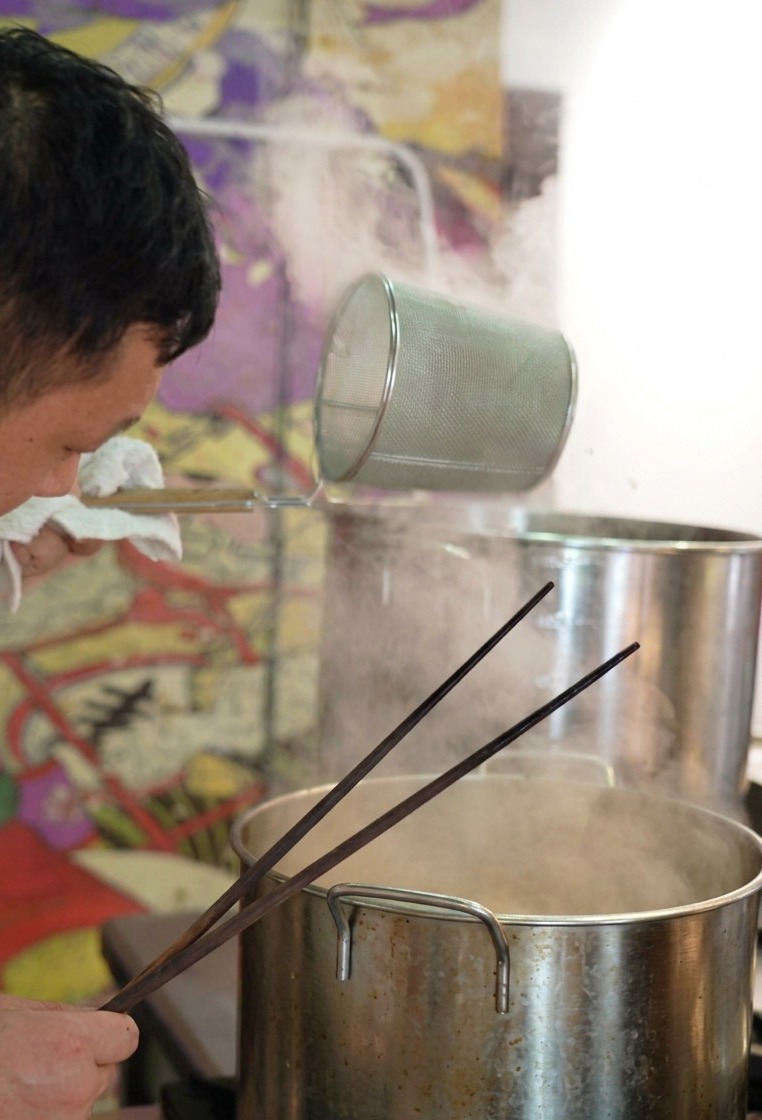 Un chef sostiene un colador cilíndrico de fideos sobre una olla humeante, sacudiéndolo para eliminar el agua de los fideos.