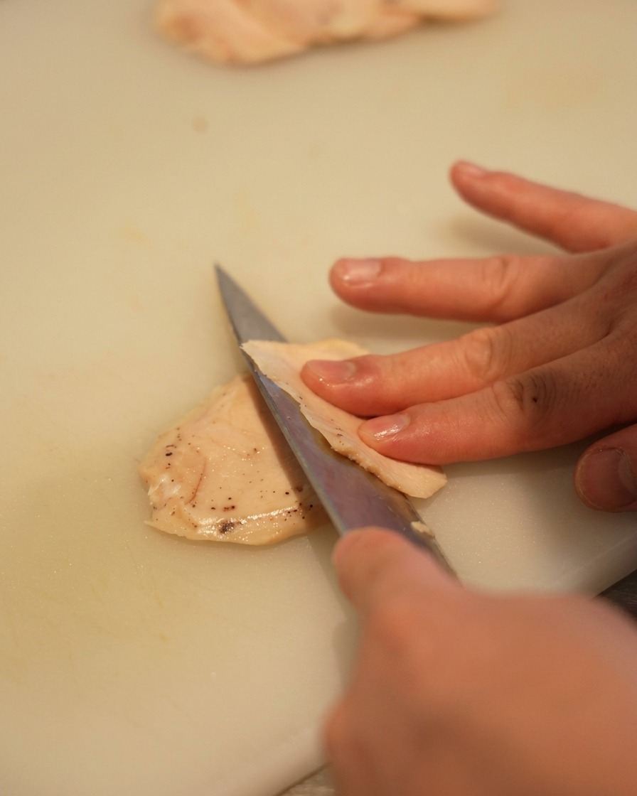 Primer plano de una mano usando un cuchillo afilado para cortar una pechuga de pollo pálida en láminas finas sobre una tabla.