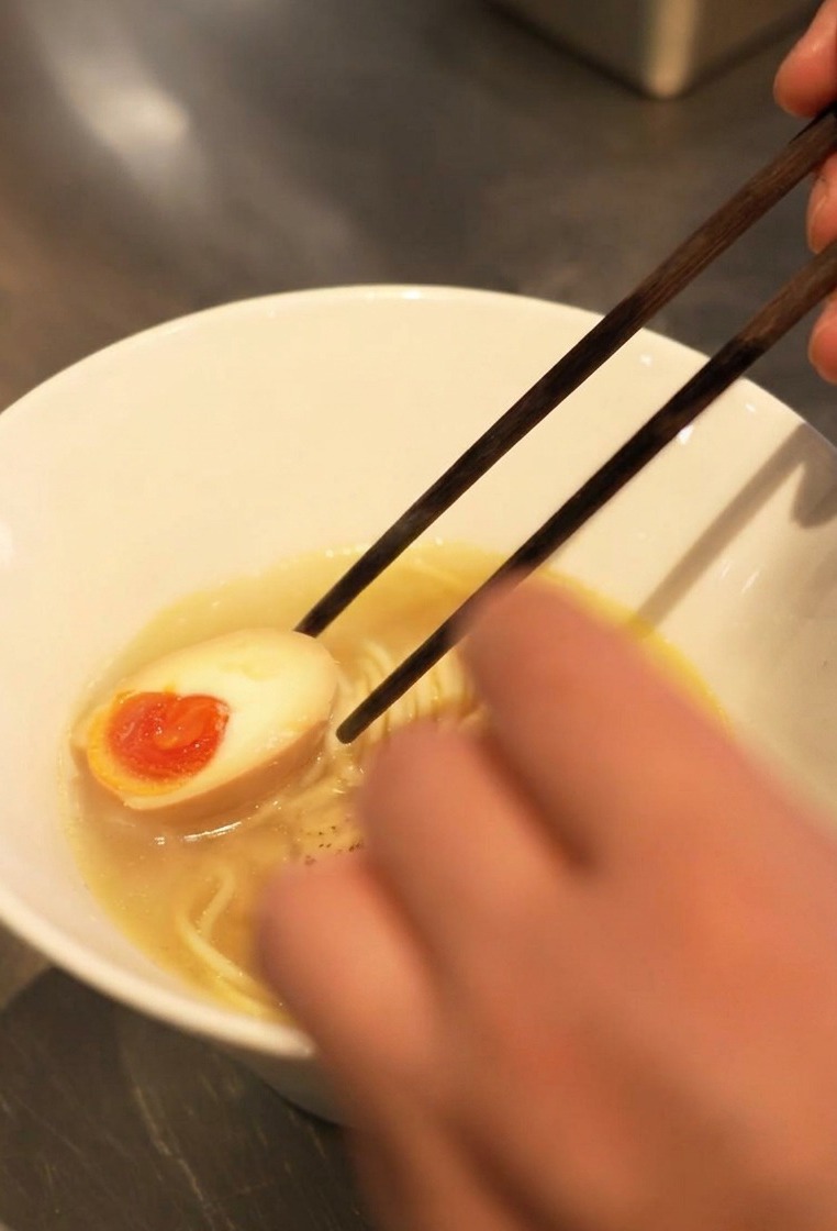 Primer plano de palillos colocando medio huevo marinado con una yema naranja brillante y líquida en un bol de ramen.