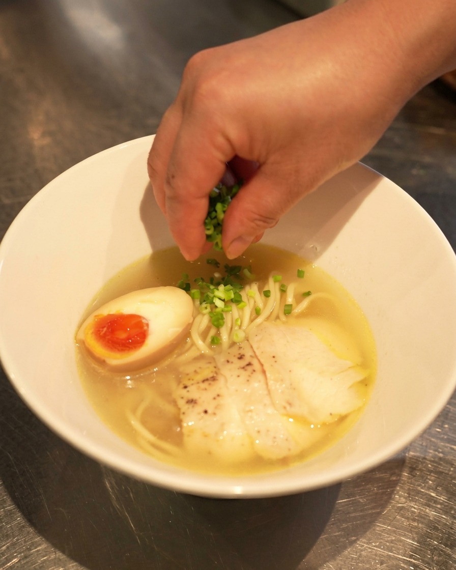 Una mano esparciendo cebolleta fresca picada sobre un bol de ramen shio terminado con huevo y pollo.