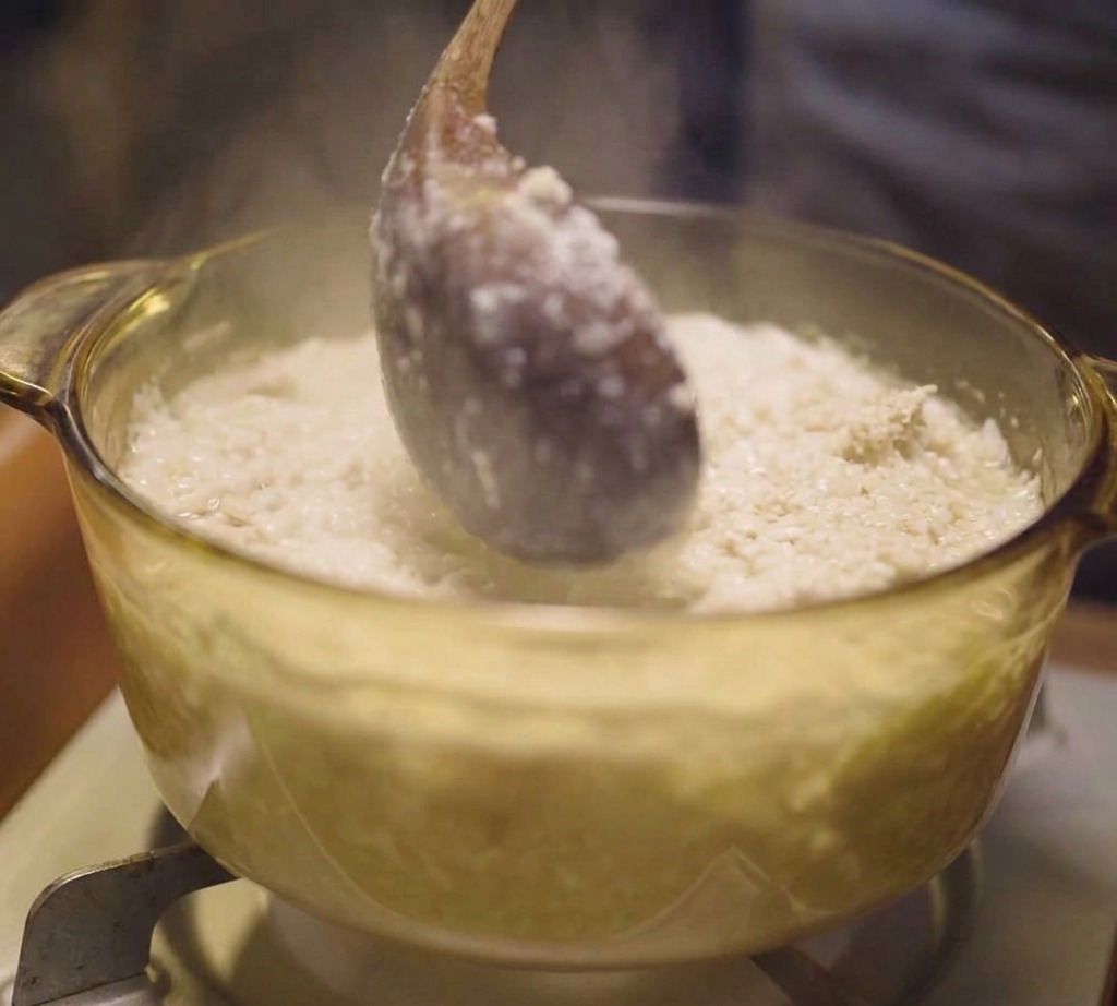 Cucharón de madera removiendo caldo de pollo con partículas de carne en una olla de vidrio.
