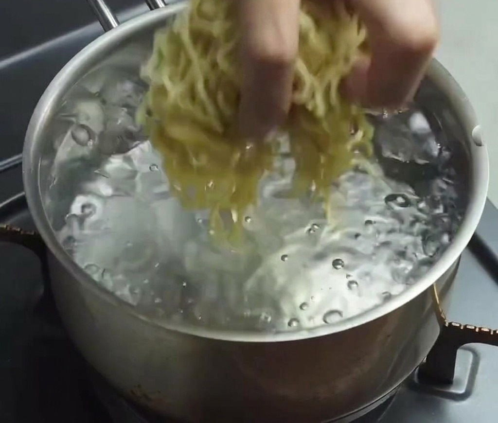Una mano soltando un fajo de fideos ramen frescos, finos y ondulados, en una olla con agua clara hirviendo a borbotones.