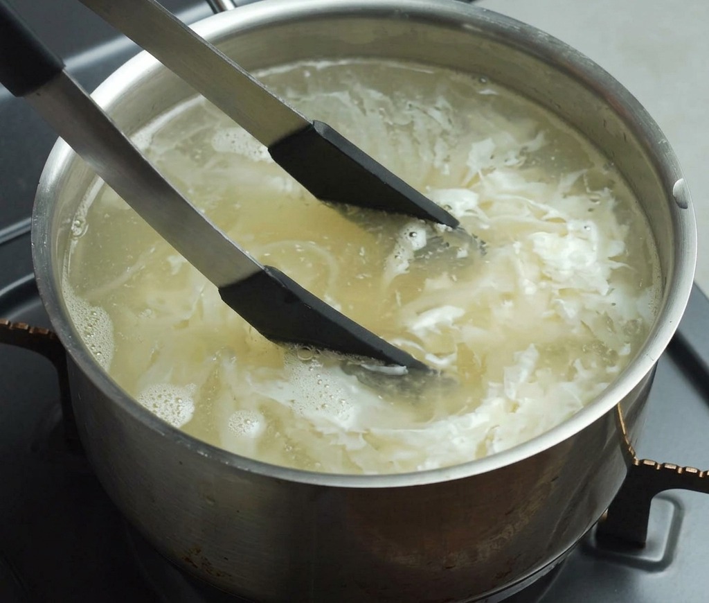 Pinzas con punta negra removiendo fideos amarillos en una olla de agua hirviendo sobre un quemador de cocina.