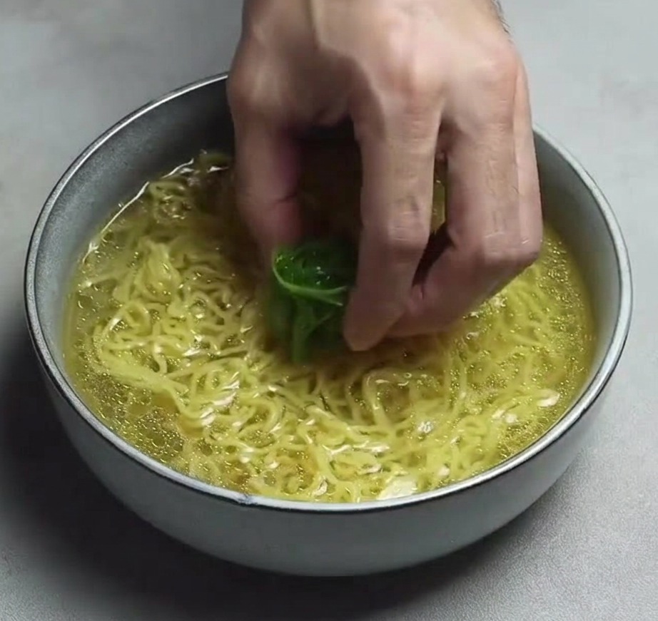 Una mano colocando un montón de tiras finas de cebolleta verde sobre los fideos ramen en un bol de caldo amarillo claro.