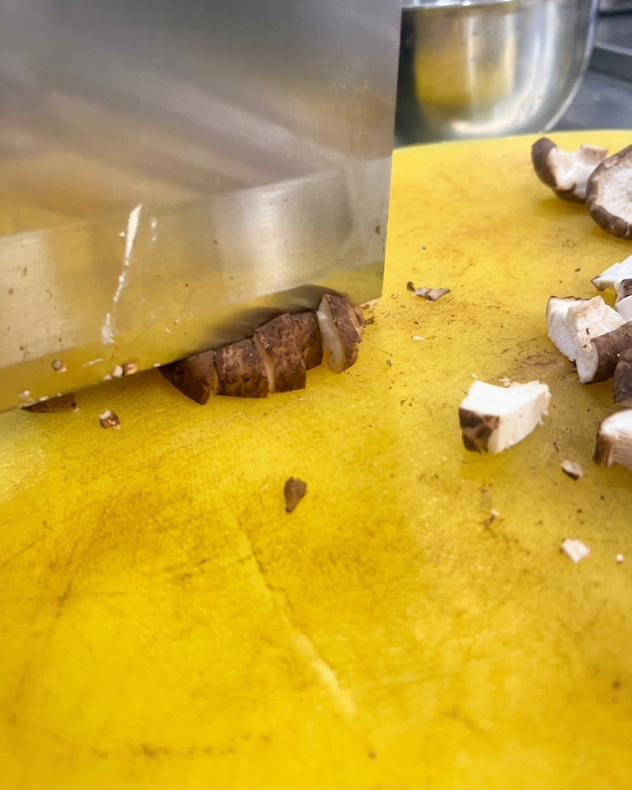 Un cuchillo de cocina picando setas shiitake en dados pequeños sobre una tabla de cortar amarilla.