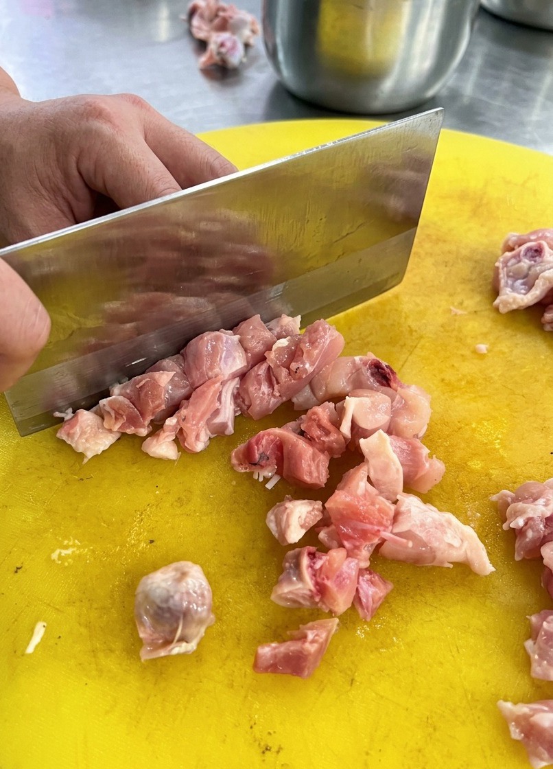 Una persona usando un hacha de cocina para trocear pollo crudo en una tabla de plástico amarilla.