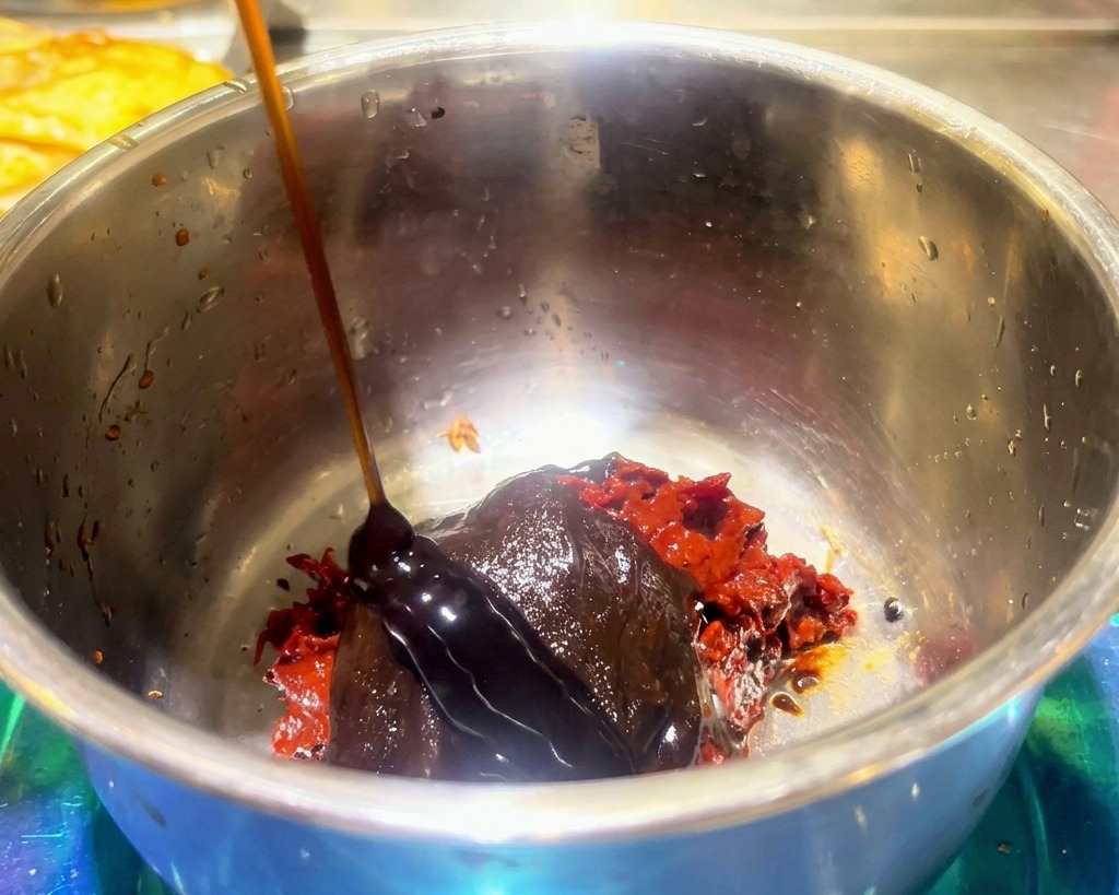 Salsa oscura espesa vertiéndose sobre pasta de judías roja y condimento de hot pot en un cuenco de acero.