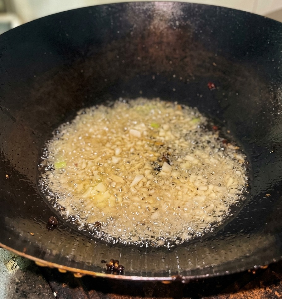 Cebolletas picadas burbujeando en aceite caliente al fondo de un wok de hierro.