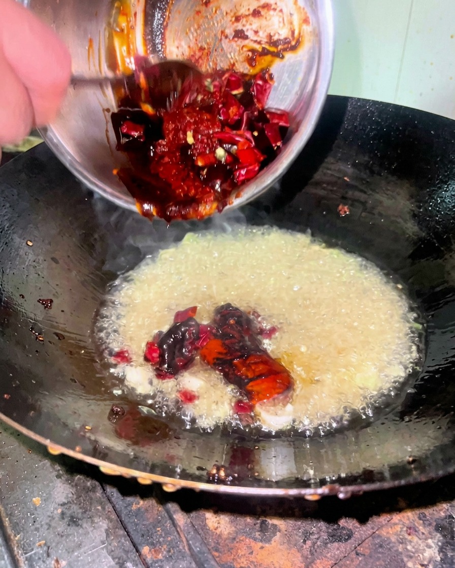 Una mezcla de salsa roja espesa cayendo de un cuenco a un wok con aceite caliente y aromáticos.