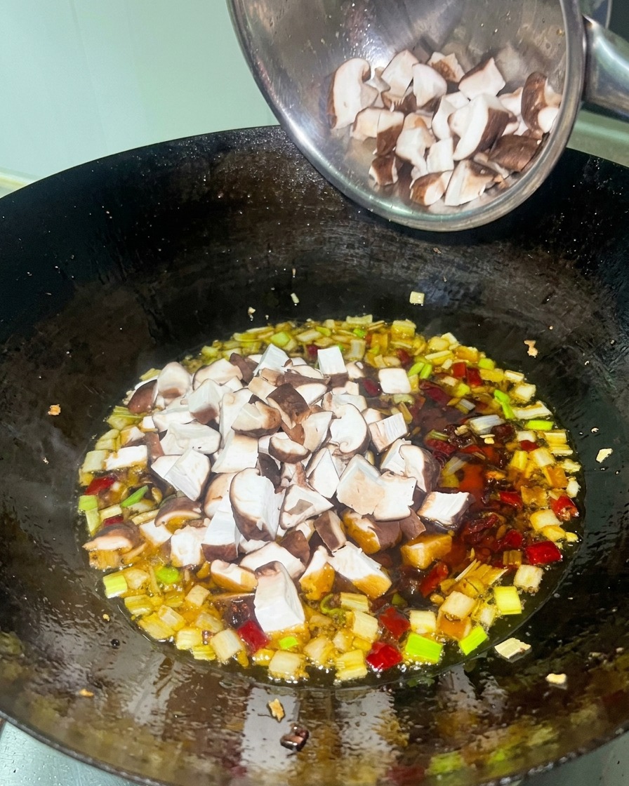 Pollo troceado y setas shiitake añadiéndose a una salsa roja densa en un wok negro.