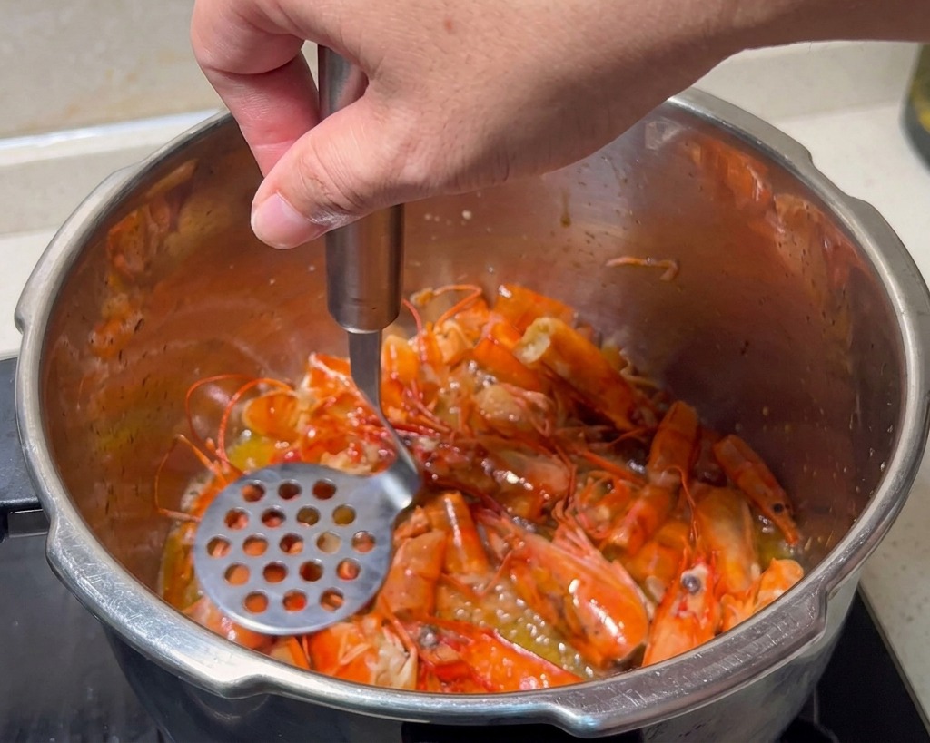 Un machacador de patatas aplastando cabezas de gamba rojas en una olla de acero inoxidable.