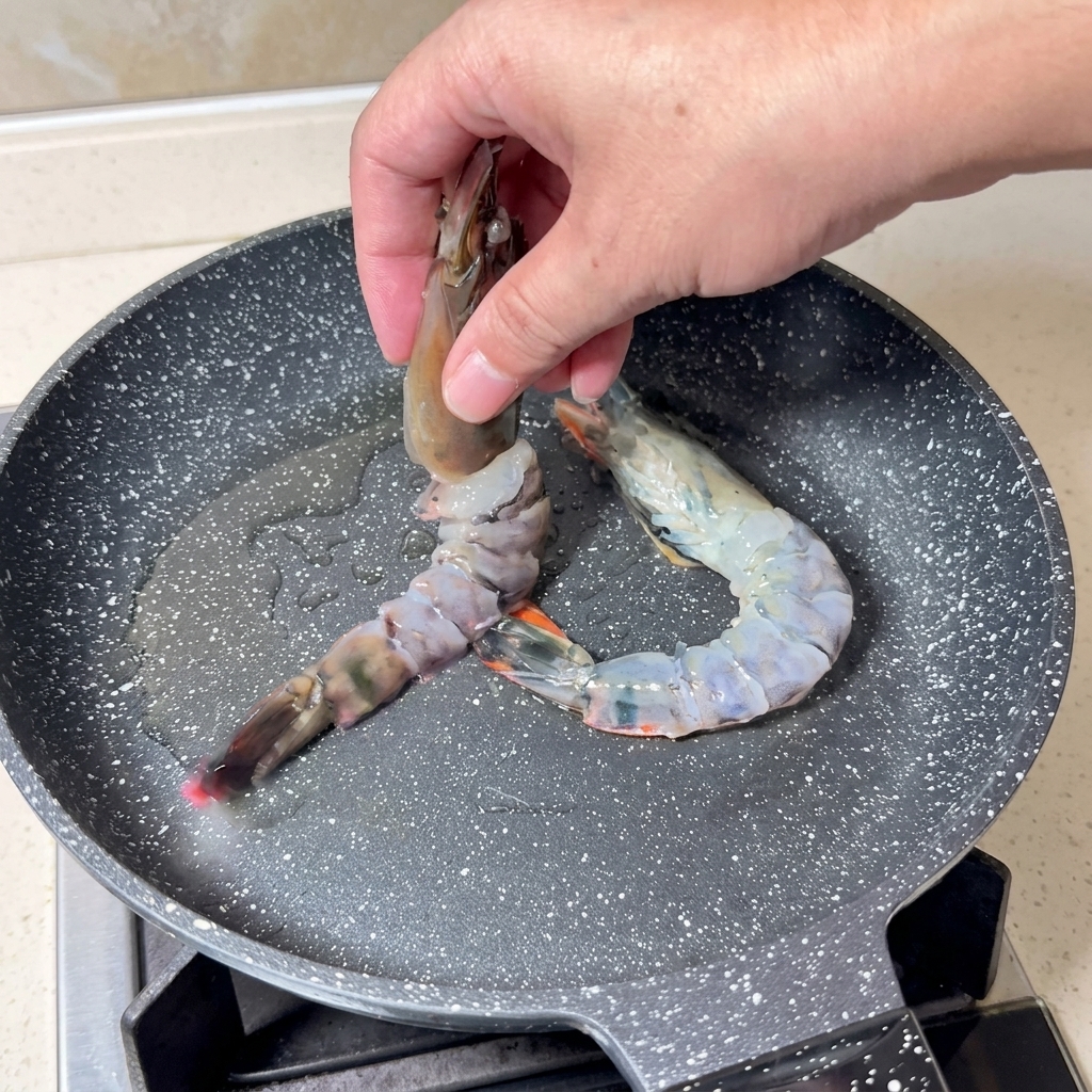 Una mano colocando langostinos crudos en una sartén caliente con aceite brillante.