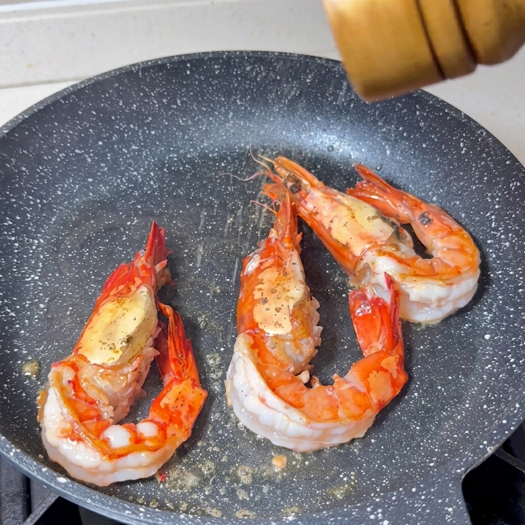 Sal y pimienta cayendo sobre tres langostinos grandes cocinándose en una sartén.
