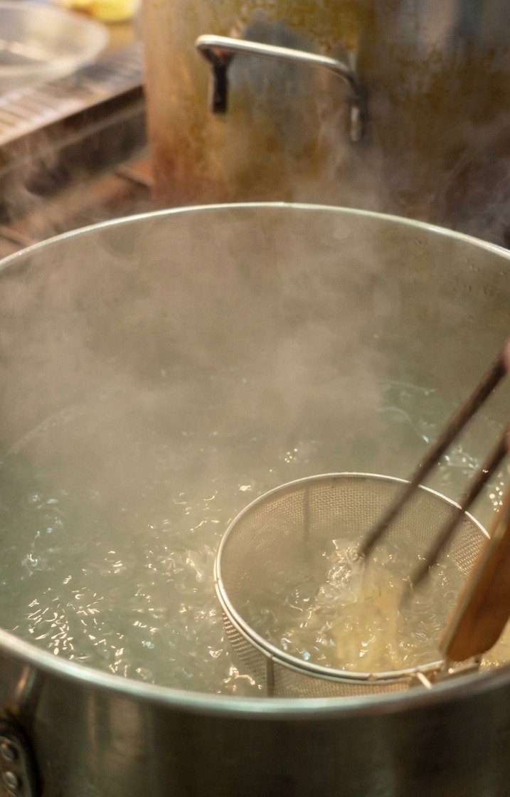 Un chef sumergiendo fideos ramen finos en una olla grande con agua hirviendo mediante un colador de malla.