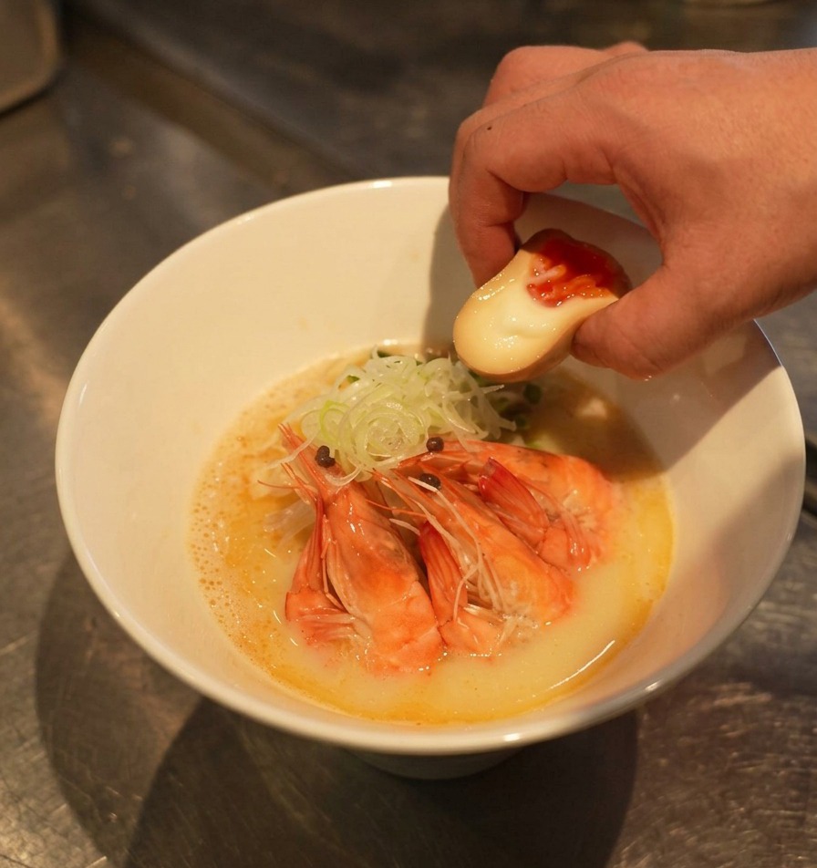 Una mano colocando medio huevo marinado con yema naranja brillante en un bol de ramen decorado con gambas rojas y cebolleta.