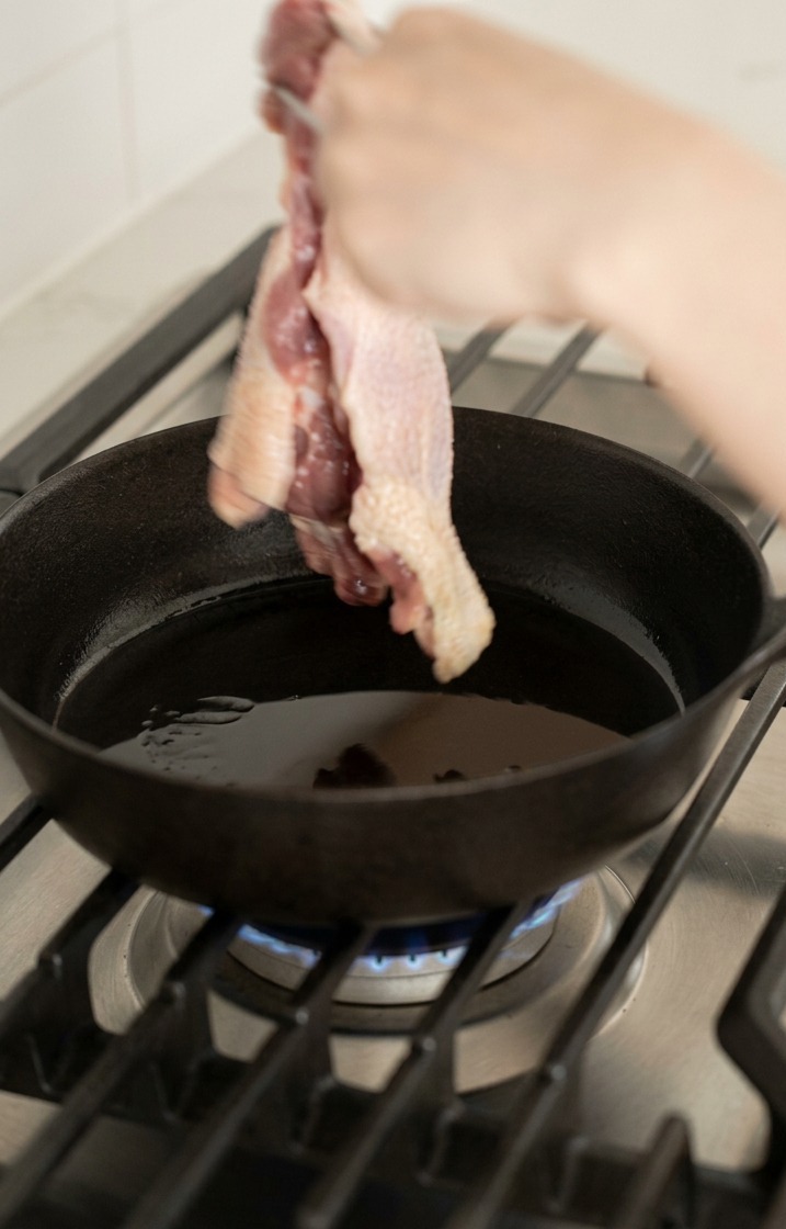 Unas pinzas colocando un muslo de pato crudo con la piel hacia abajo en una sartén de hierro fundido caliente con aceite.