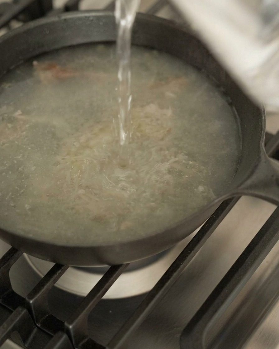 Agua vertiéndose en una sartén oscura con jugos de carne y aromáticos sobre un fuego de gas.